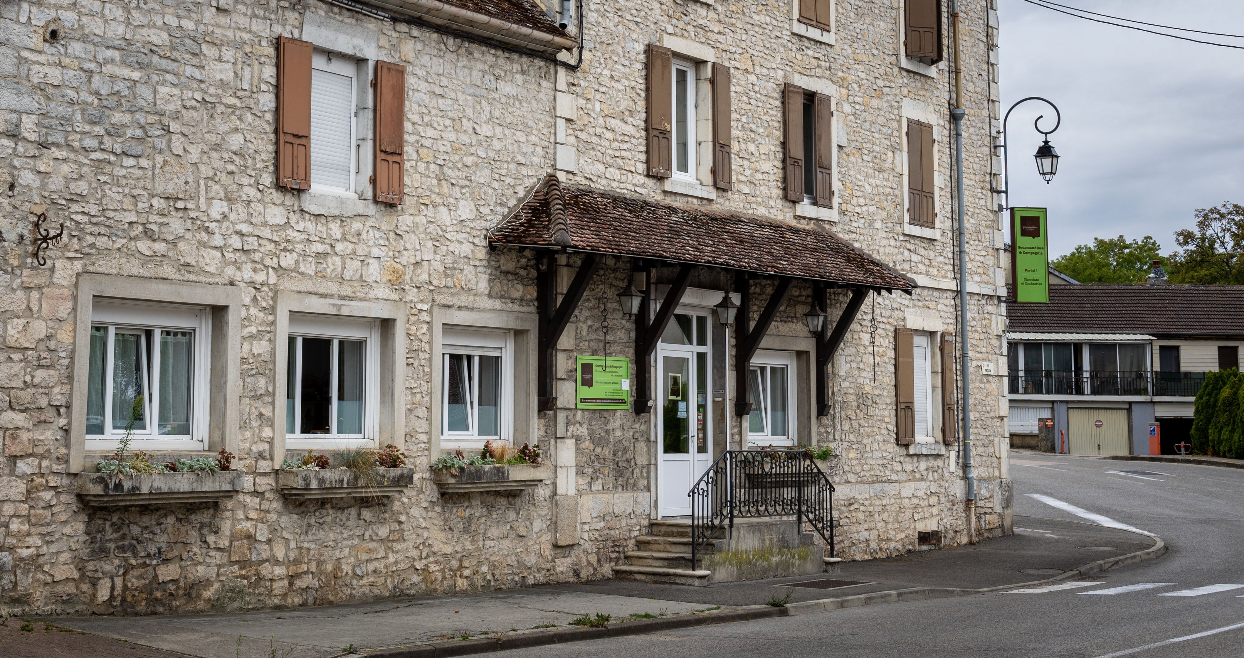 La façade en pierre de l’atelier Gourmandises & Cie, au 5 rue Progin à Champagnole, avec son auvent de tuiles et son enseigne verte au coin de la rue.